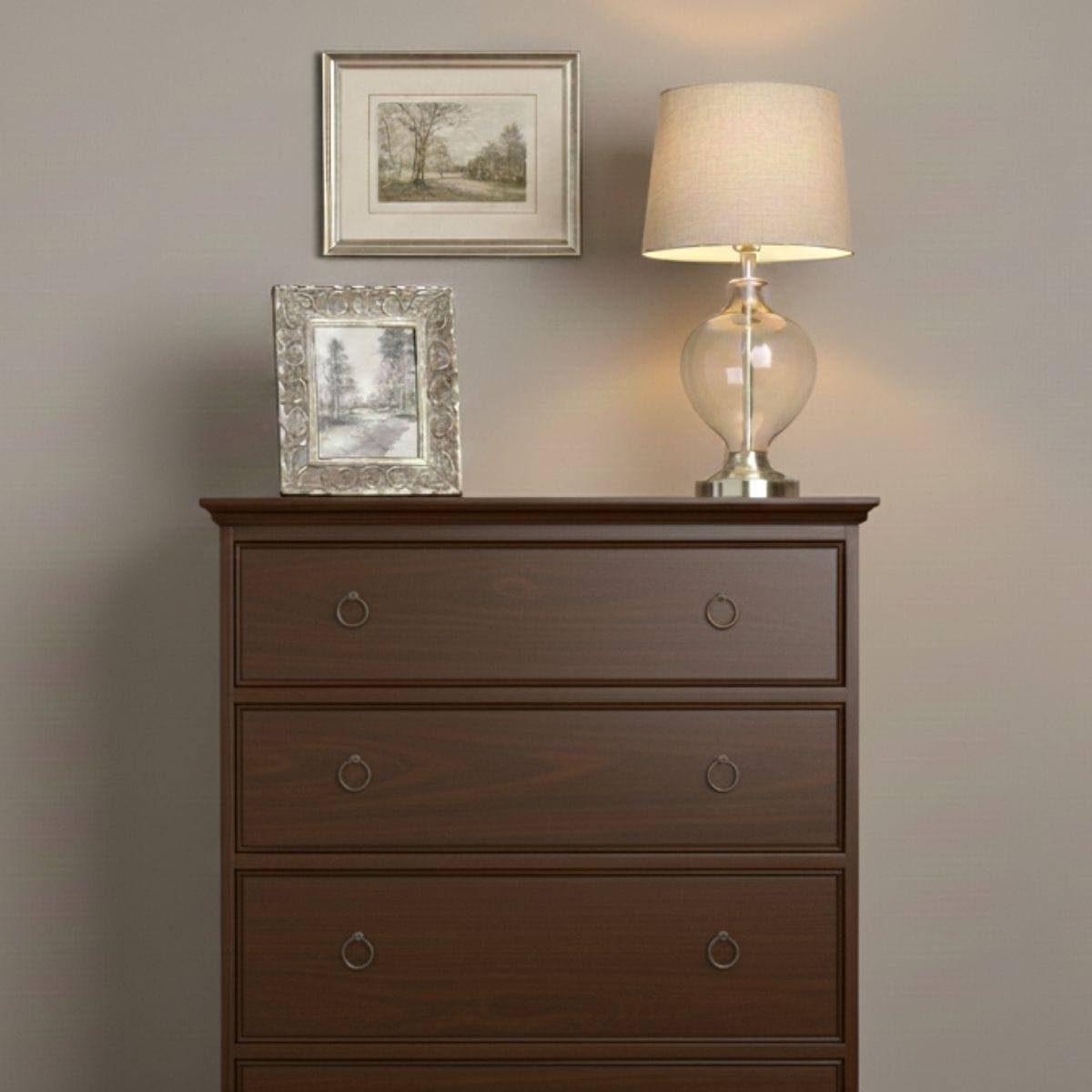A classic four-drawer chest in a rich, dark antique walnut finish. The chest is set against a neutral, sand-colored wall (#E3D5B8). On top of the chest sits an ornate silver picture frame containing a landscape sketch, a blue and white Chinoiserie ginger jar, and a lamp with a hand-blown clear glass base, silver hardware, and a light linen shade. The lighting is warm and soft, creating a sophisticated and cozy atmosphere.