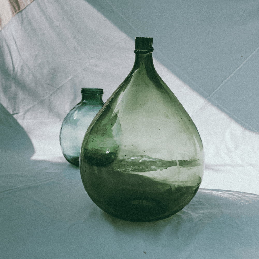 A green glass vase with a bulbous shape placed on a rustic wooden table, styled with neutral ceramics in the background. Natural light illuminates the glass, highlighting its translucent texture and sculptural form.